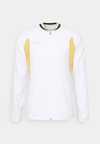 Weißer Sportjacke mit langen Ärmeln, Frontreißverschluss, goldenen Seitenteilen, goldenem Nike-Logo auf der linken Brust und Kragen mit schwarzen und goldenen Akzenten.