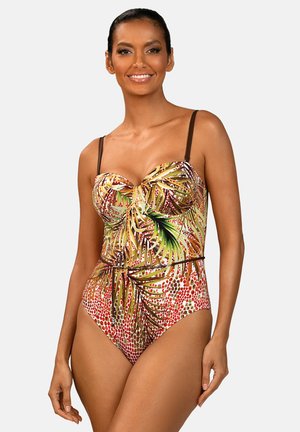 Kobieta w jednoczęściowym kostiumie kąpielowym z tropikalnym wzorem liści i kropek, uśmiechająca się i stojąca na jednolitym tle.