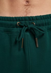 Grønne sweatpants med snore i taljen, med metaløjer og snorelåse, lavet af et blødt, tekstureret stof.