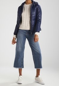 Person in einer marineblauen Daunenjacke über einem cremefarbenen Strickpullover, mit blauen, gekürzten weit geschnittenen Jeans und weißen Sneakers, stehend vor einem weißen Hintergrund.