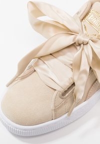 Sneaker in suede beige con ampie stringhe in raso, suola in gomma bianca e un accento dorato del logo sulla linguetta.
