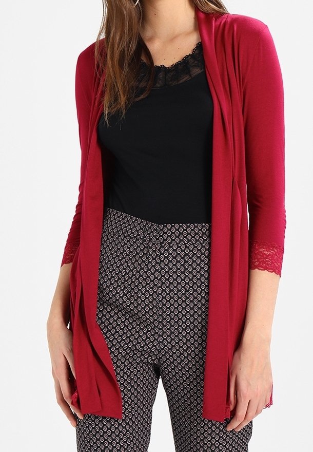 Femme portant un cardigan rouge ouvert avec une bordure en dentelle sur un haut noir bordé de dentelle et un pantalon à motifs noir et blanc, debout les mains détendues.