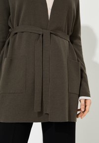 Cardigan vert olive en tissu tricoté doux, doté d'une taille ceinturée, de deux poches avant et de poignets clairs contrastants.