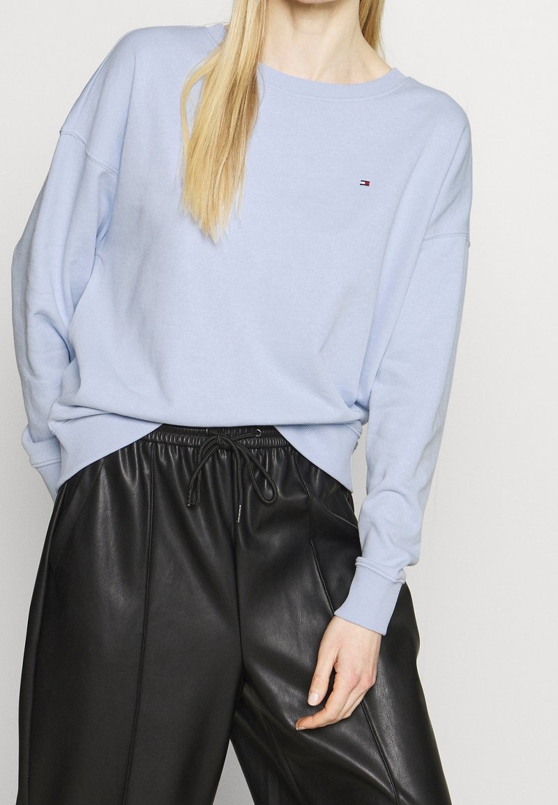 Personne portant un sweat-shirt à manches longues bleu clair avec un petit logo et un pantalon en cuir noir à cordon, visible du cou jusqu'au milieu de la cuisse.