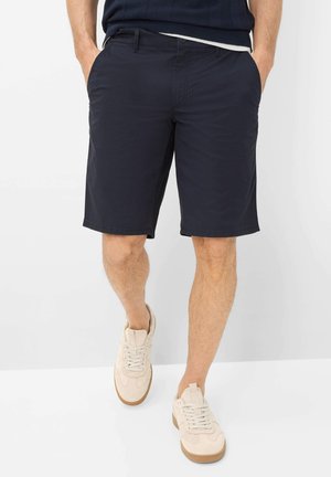 Mand iført marineblå knælange shorts og beige sneakers, stående med hænderne i lommerne mod en hvid baggrund.