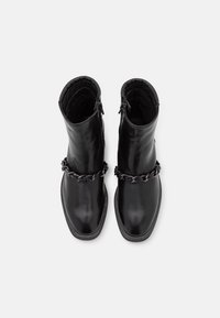 Par de botas de cuero negras hasta el tobillo con puntas cuadradas y cadenas de metal oscuro decorativas en la parte frontal, mostradas desde arriba sobre un fondo blanco.