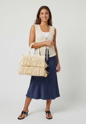 Jonge vrouw in mouwloze crème gebreide top, donkerblauwe satijnen midi rok, zwarte sandalen met hak, grote beige franjes tas vasthoudend, glimlachend.