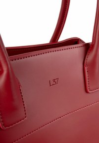 Raudona odinė rankinė su lygiu paviršiumi, stačiakampio formos, turinti iškilų "L37" detalę ir tvirtas dviem rankenomis.