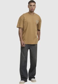 Brązowa bawełniana koszulka z krótkimi rękawami i okrągłym dekoltem, z graficznym wzorem po lewej stronie klatki piersiowej; połączona z czarnymi spodniami o szerokich nogawkach.