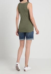 Kvinde iført en olivengrøn ærmeløs top, opsmøgede denimshorts og hvide sneakers, stående med ryggen til beskueren.