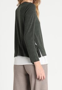 Pull vert foncé à manches longues, avec un détail de lien sur le côté, et une chemise blanche superposée en dessous, associé à un pantalon à carreaux.