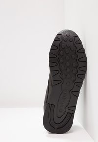 A sola do ténis preto apresenta um piso de borracha texturizada com padrões circulares, lados sulcados e uma impressão de logótipo. O design é leve e durável.