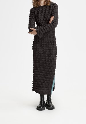 Woman wearing a long textured black dress with puffed sleeves and black chunky boots, standing with arms crossed against white background.