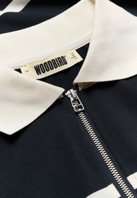 Vêtement noir avec col blanc et fermeture éclair argentée, affichant l'étiquette de marque "WOODBIRD" à l'intérieur du col.