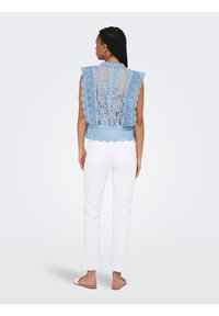 Femme aux cheveux tressés portant un haut en dentelle bleu sans manches et un pantalon blanc, debout de dos sur un fond clair uni.