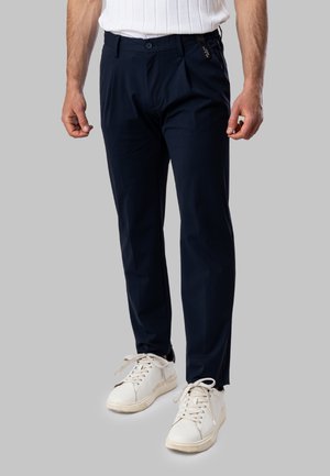 Uomo che indossa pantaloni slim fit blu navy e sneaker bianche, in piedi davanti a uno sfondo grigio uniforme.