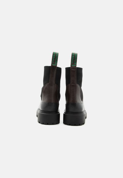 Ζευγάρι μαύρα και σκούρα καφέ δερμάτινα μποτάκια με χοντρές σόλες, ορατά από πίσω, με πράσινες ετικέτες τραβήγματος με λογότυπο.