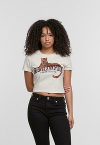 Kremasta skraćena majica s grafikom tigra i natpisom "True Religion Brand Jeans". Kratki rukavi i prilagođeni dizajn. Crne traperice visokog struka.