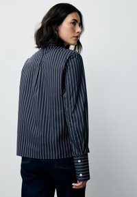 Marineblauw overhemd met witte verticale strepen, manchetten met knopen en een achterplooi voor extra structuur. Katoenachtige textuur met een getailleerde pasvorm.