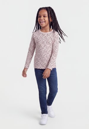 Lachend meisje dat loopt, draagt een lichtroze shirt met lange mouwen met zwarte stippen, donkerblauwe spijkerbroek en witte sneakers.