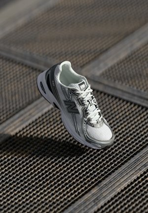 Scarpa da corsa New Balance bianca e grigia con lacci, posizionata su una superficie di grata metallica con un motivo testurizzato.