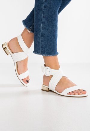 Des pieds dans des sandales ouvertes blanches à talons blocs dorés et brides de cheville, associées à un jean bleu avec ourlet effiloché sur un sol blanc.