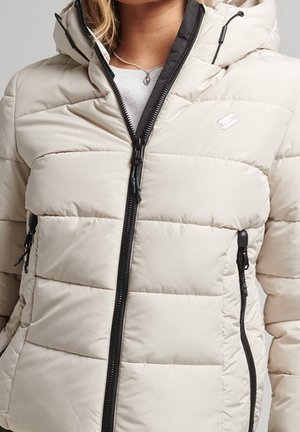 Kobieta ubrana w jasnobeżową pikowaną kurtkę puchową z czarnym zamkiem i kapturem, częściowo rozpiętą na jasny top z zawieszką na szyi.