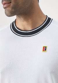 Chemise à manches courtes en tissu blanc perforé avec un col noir à côtes comportant des rayures blanches. Petit logo multicolore sur la poitrine.