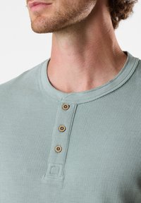 Vaaleanvihreä ribattu henley-paita, jossa on kolme puista nappia ja pyöreä kaula-aukko. Paita on valmistettu tekstuurista kankaasta ja siinä on pieni neliömäinen yksityiskohta.