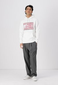 Hvit hettegenser med en rosa grafisk logo, parret med grå bukser og hvite sneakers. Enkelt design, avslappet passform og glatt tekstur.