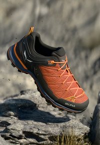 Zwart en oranje sportschoen met een gestructureerde mesh bovenkant, versterkte neus en een opvallend vetersysteem, geschikt voor buitensportactiviteiten.