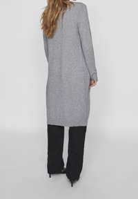 Cardigan long en maille grise avec ourlet et poignets côtelés, présentant une coupe ample et une longueur mi-longue. Porté sur un pantalon noir.