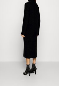 Svart långärmad klänning med hög hals, tillverkad av ett texturerat tyg; kombineras med svarta klackar och mönstrade ankelsockor.
