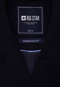 Cuello de chaqueta de mezclilla negra con una etiqueta que dice "BIG STAR", talla M, y "CALIDAD PREMIUM". Textura suave con costuras mínimas.