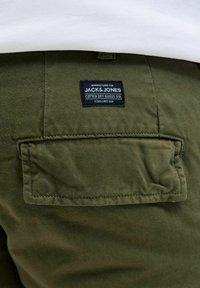 Pantalones de algodón verde oliva con un bolsillo trasero. Presenta una etiqueta negra que dice "JACK & JONES COTTON DRY GOODS DIV." Fundada en 1990.