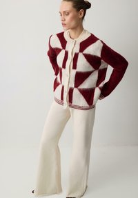 Sweter kardigan z geometrycznym wzorem w kolorze bordowym i kremowym, okrągłym dekoltem oraz zapięciem na guziki. W zestawie z kremowymi spódnicami w kształcie dzwonu.