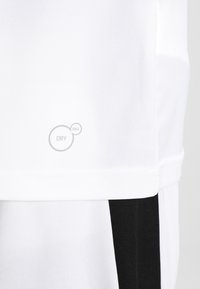 Vit tyg med "DRY CELL"-logotyp nära fållen, kombinerat med svart och vitt strukturerat tyg nedanför, vilket antyder sportklädesmaterial.