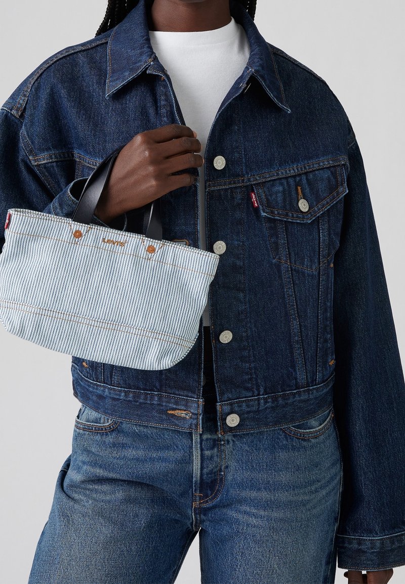 Person trägt eine dunkelblaue Jeansjacke und Jeans, hält eine kleine hellblaue gestreifte Tragetasche mit schwarzen Griffen.