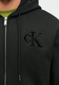 Μαύρο φούτερ με φερμουάρ από υφασμάτινη δομή, με κεντημένο λογότυπο CK στο στήθος και ασημί λεπτομέρεια στο φερμουάρ.