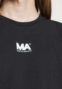 Nærbillede af en person iført en sort skjorte med hvidt "MA Martin Asbjørn" logo trykt tæt på kraven.