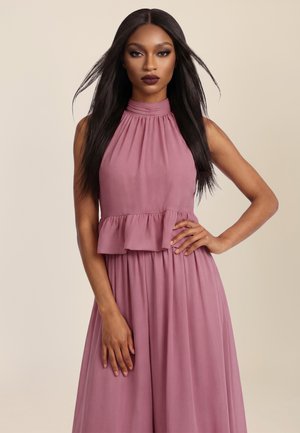 Frau mit langen schwarzen Haaren trägt ärmelloses mauvefarbenes Kleid mit hohem Kragen und geraffter Taille, steht vor beigem Hintergrund.