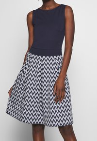 Sukienka bez rękawów o granatowej górze i rozkloszowanej spódnicy z wzorem w zygzaki w granacie i bieli, wykonana z gładkiego materiału.