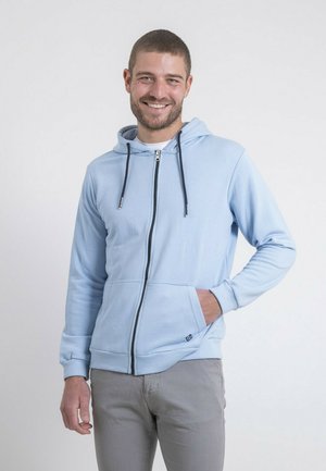 Homme souriant portant un sweat à capuche bleu clair zippé, les mains dans les poches, debout devant un fond clair uni.