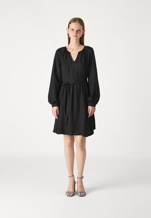 Robe noire avec un décolleté en V, de longues manches bouffantes et une taille cintrée. Fabriquée en tissu texturé, se terminant au-dessus du genou. Associée à des sandales à talons bleues.