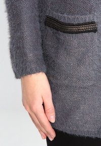 Detailní záběr na ruku vedle huňatého šedého pleteného svetru s texturovaným vzorem a malým dekorativním černým lemem na kapse.