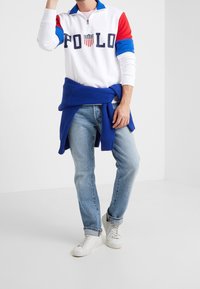 Bílá polo košile s dlouhým rukávem a červenými a modrými rukávy, se zipovým límcem a logem "POLO". Oblečení doplňují světle modré džíny a bílé tenisky.