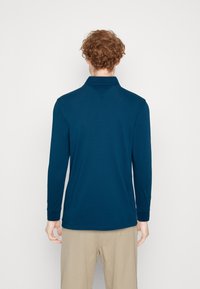 Marinblå långärmad polo med krage, tillverkad av ett mjukt tyg, med rak fåll och åtsittande mudd, parat med ljusa byxor.