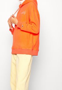 Osoba w pomarańczowej bluzie z kapturem z białym logo na piersi i rękawie, poprawiająca sznurki, w zestawieniu z jasnobeżowymi spodniami z kordurowej tkaniny.