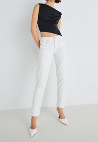 Černý top bez rukávů s twistovaným designem, doplněný bílými džíny s rovnými nohavicemi a bílými lodičkami s špičatou špičkou na neutrálním pozadí.