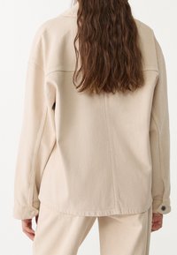 Beige denim jacket with a relaxed fit, featuring a visible seam down the center, buttoned cuffs, and soft texture.